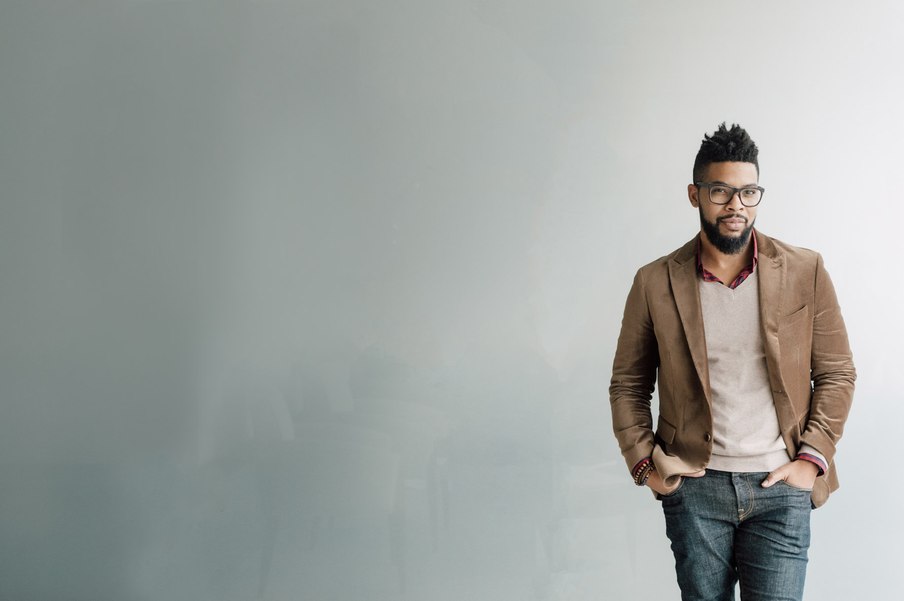 A stylish black man in a tan jacket and jeans posing against a plain gray background for business photography.