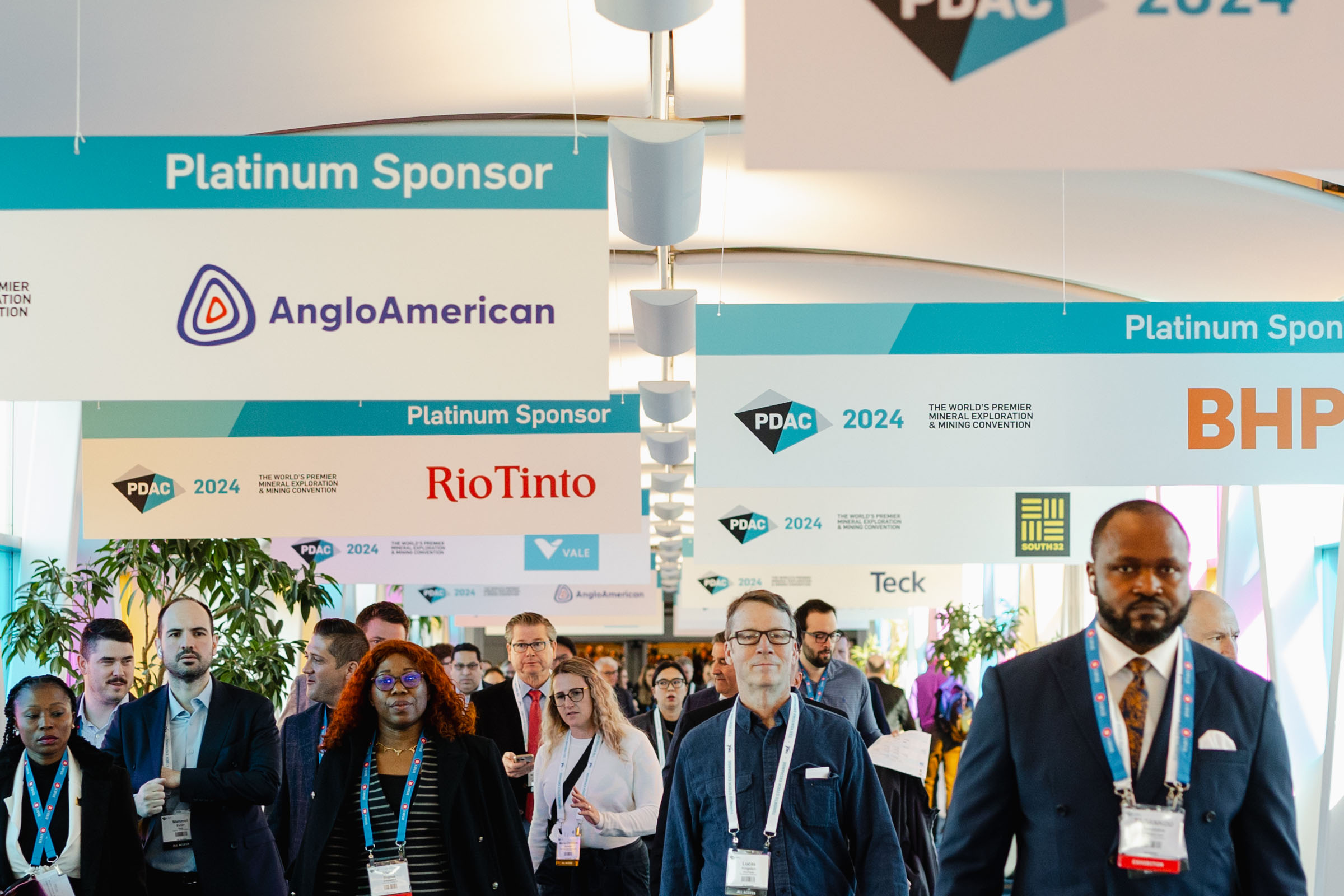 Attendees walking through a convention hall with sponsor banners for a corporate photography event, prominently featuring mining companies like Anglo American and Rio Tinto.