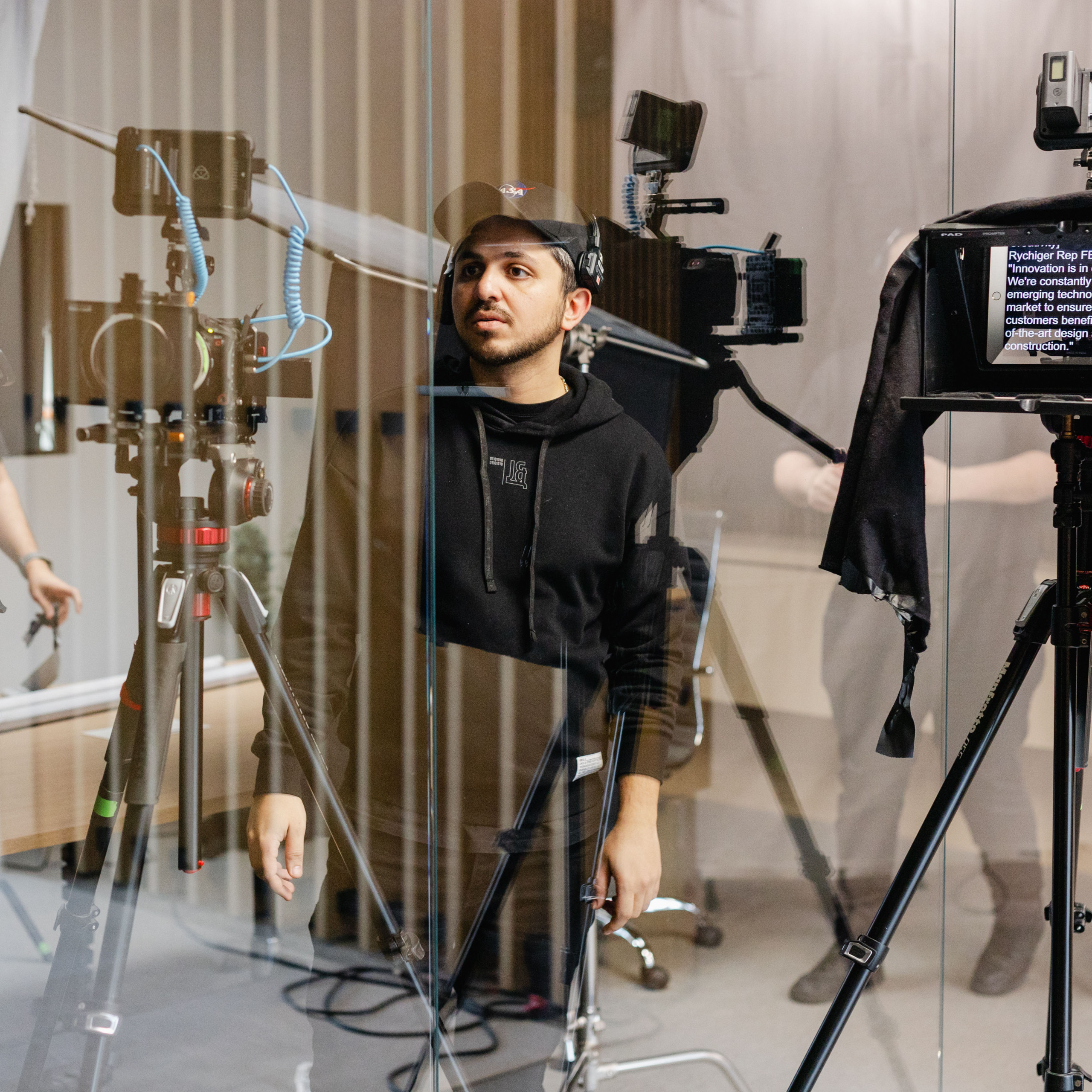 A man in a black hoodie and cap stands among camera equipment and lights in a studio setting, viewed through a glass panel, capturing the essence of corporate photography.