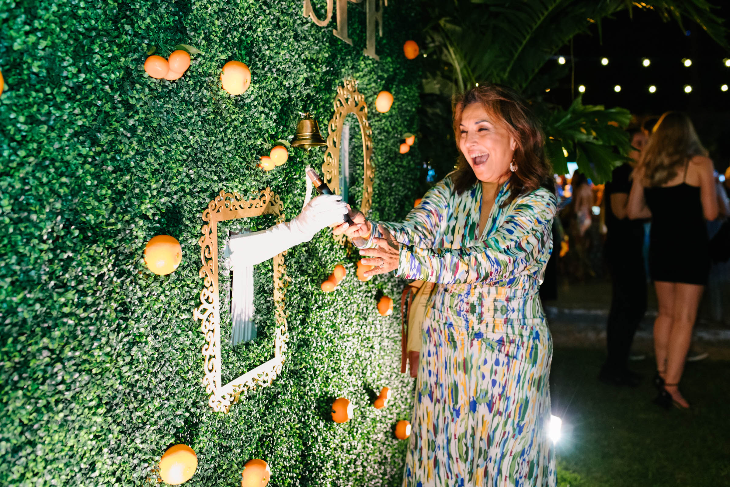 A woman in a patterned dress poses for corporate photography as she interacts with a green decorative wall adorned with oranges and framed cutouts.